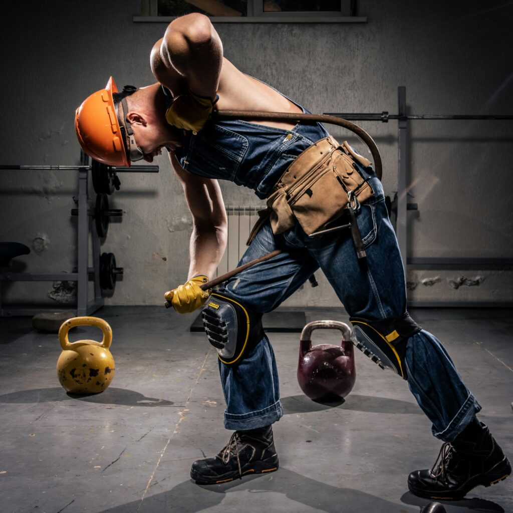 Man in construction gear bending with kettlebells indoors, showcasing strength and fitness.