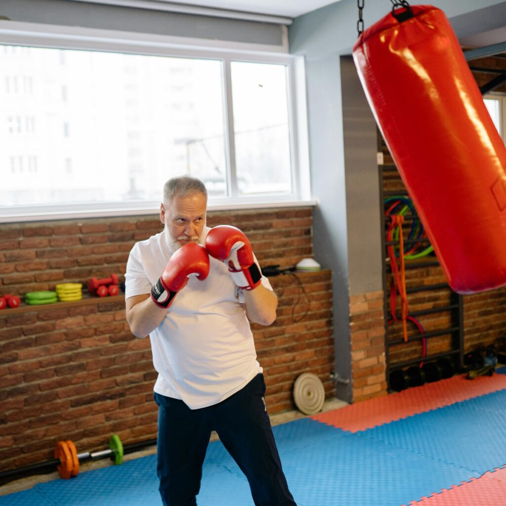 Elderly man practicing boxing indoors with red gloves and heavy bag.